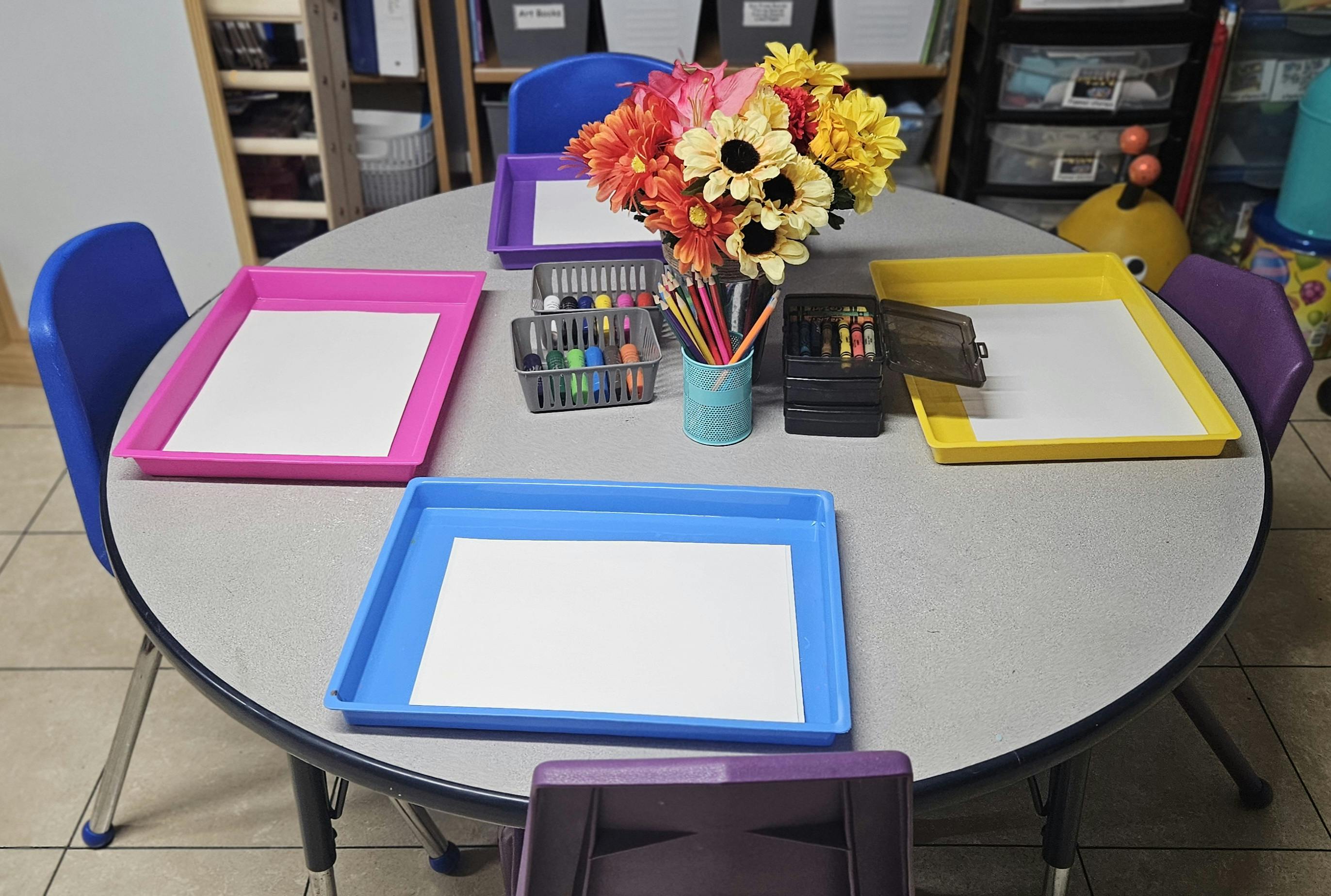 Round table with colorful trays, paper, art supplies, and a flower bouquet
