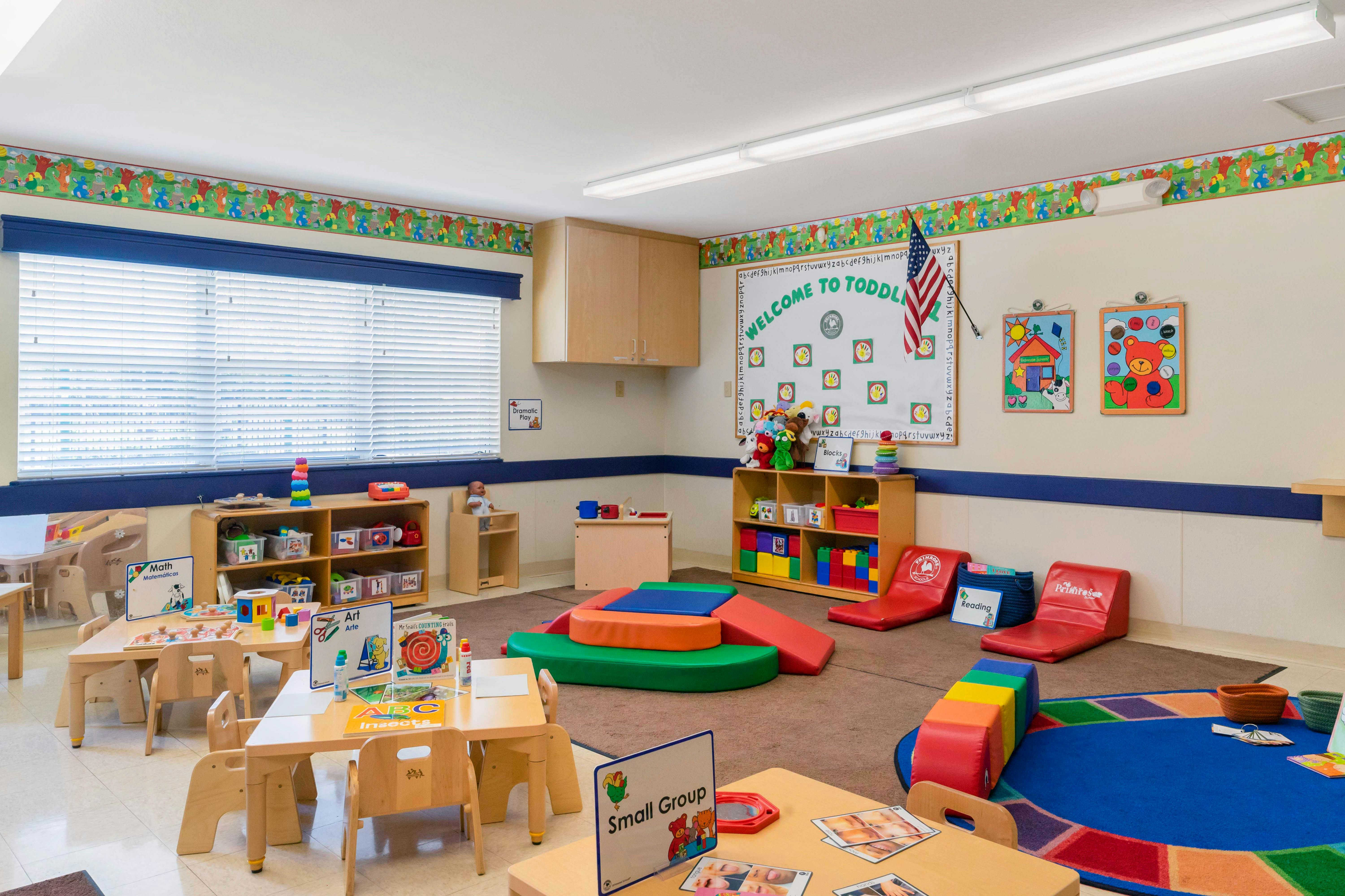 A colorful empty classroom with small tables, chairs, play mats, and a welcome sign