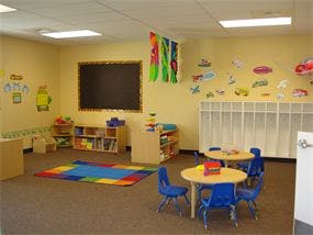 Empty classroom with small tables and chairs, rug, and chalkboard