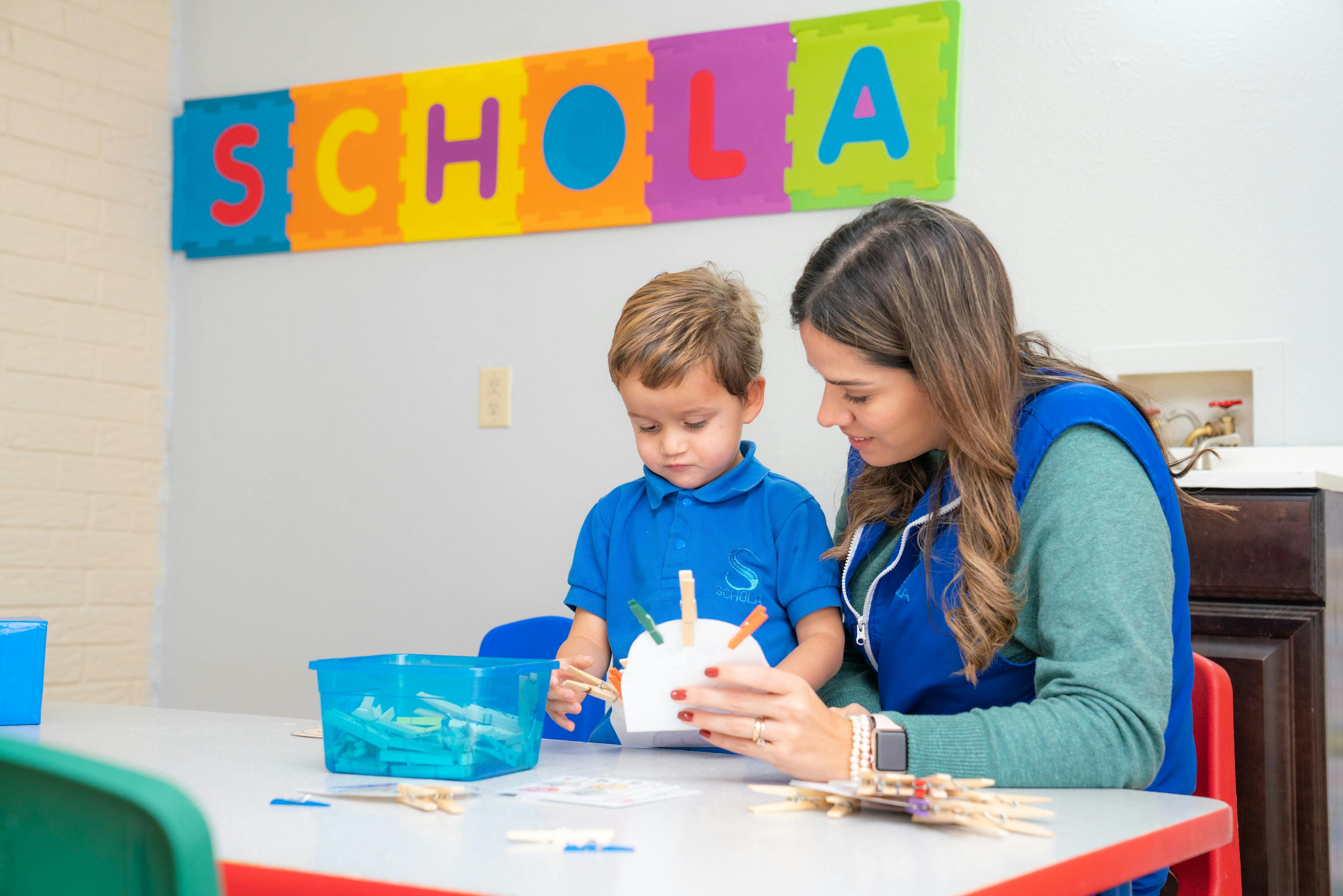 Adult and child at a table with craft supplies, sign says SCHOLA