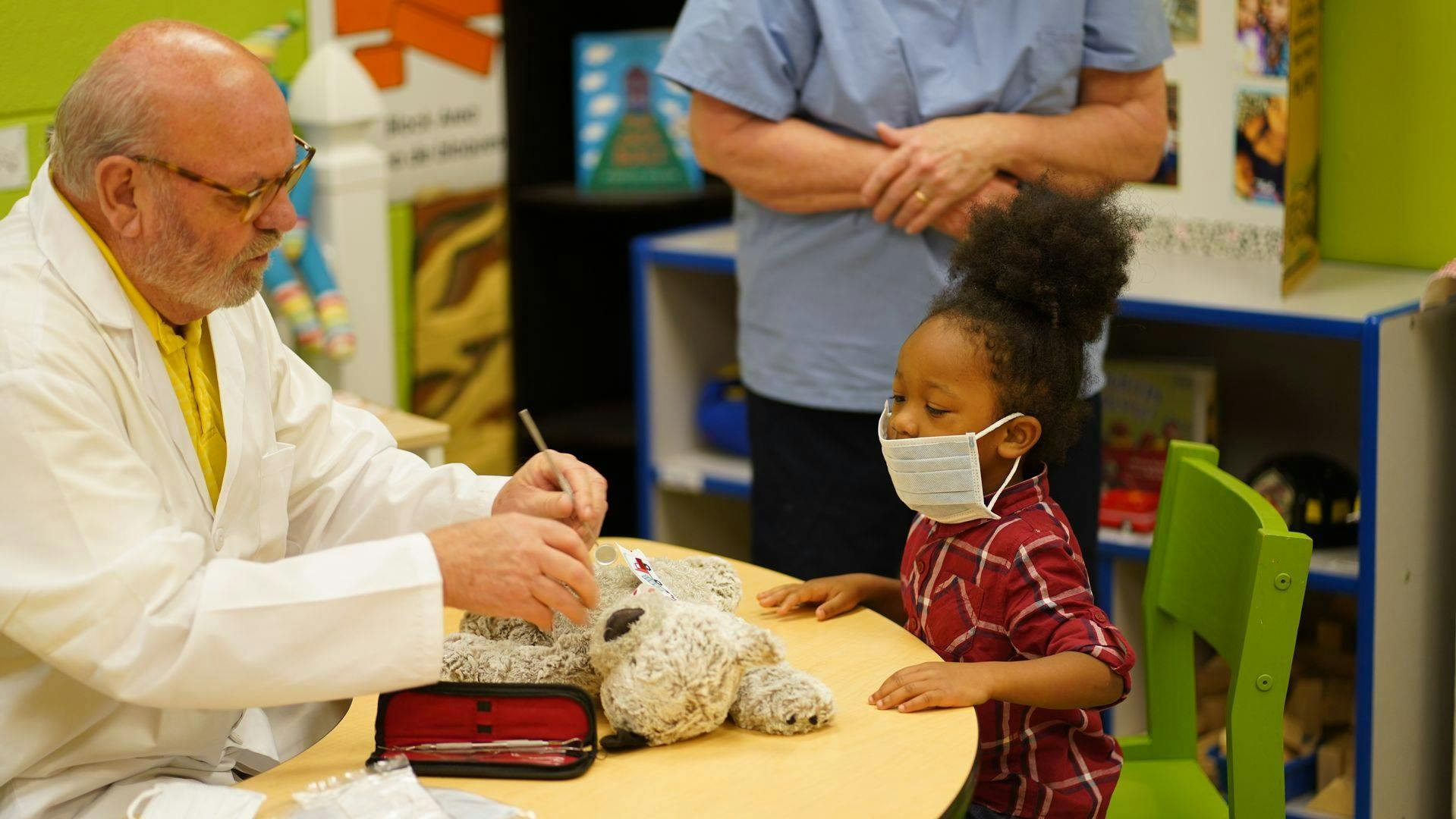 Person in lab coat working on stuffed animal with child in mask watching
