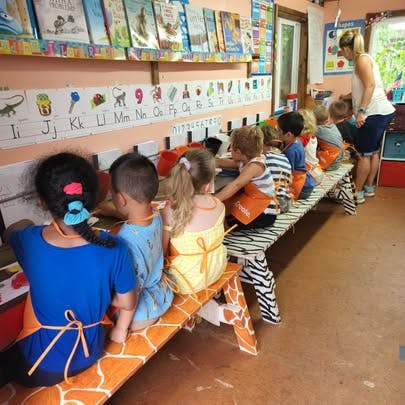 Children sitting at a long table in a classroom with an adult standing nearby