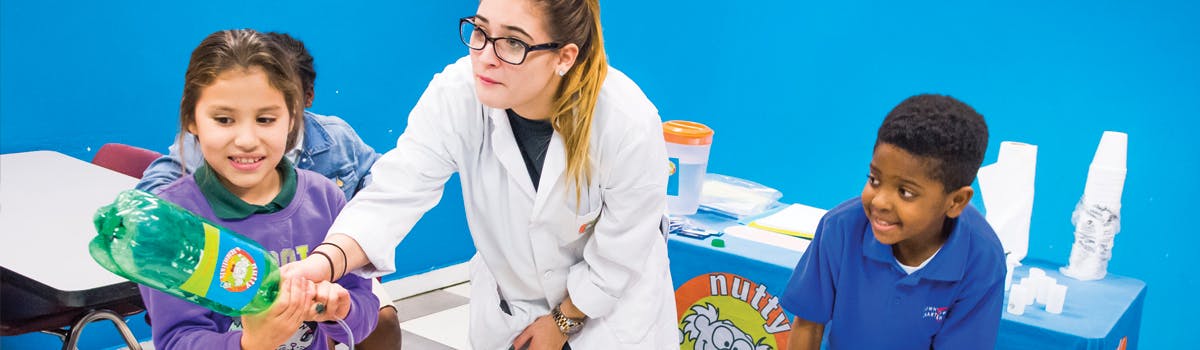 Person in lab coat helping child with a plastic bottle, another child watching