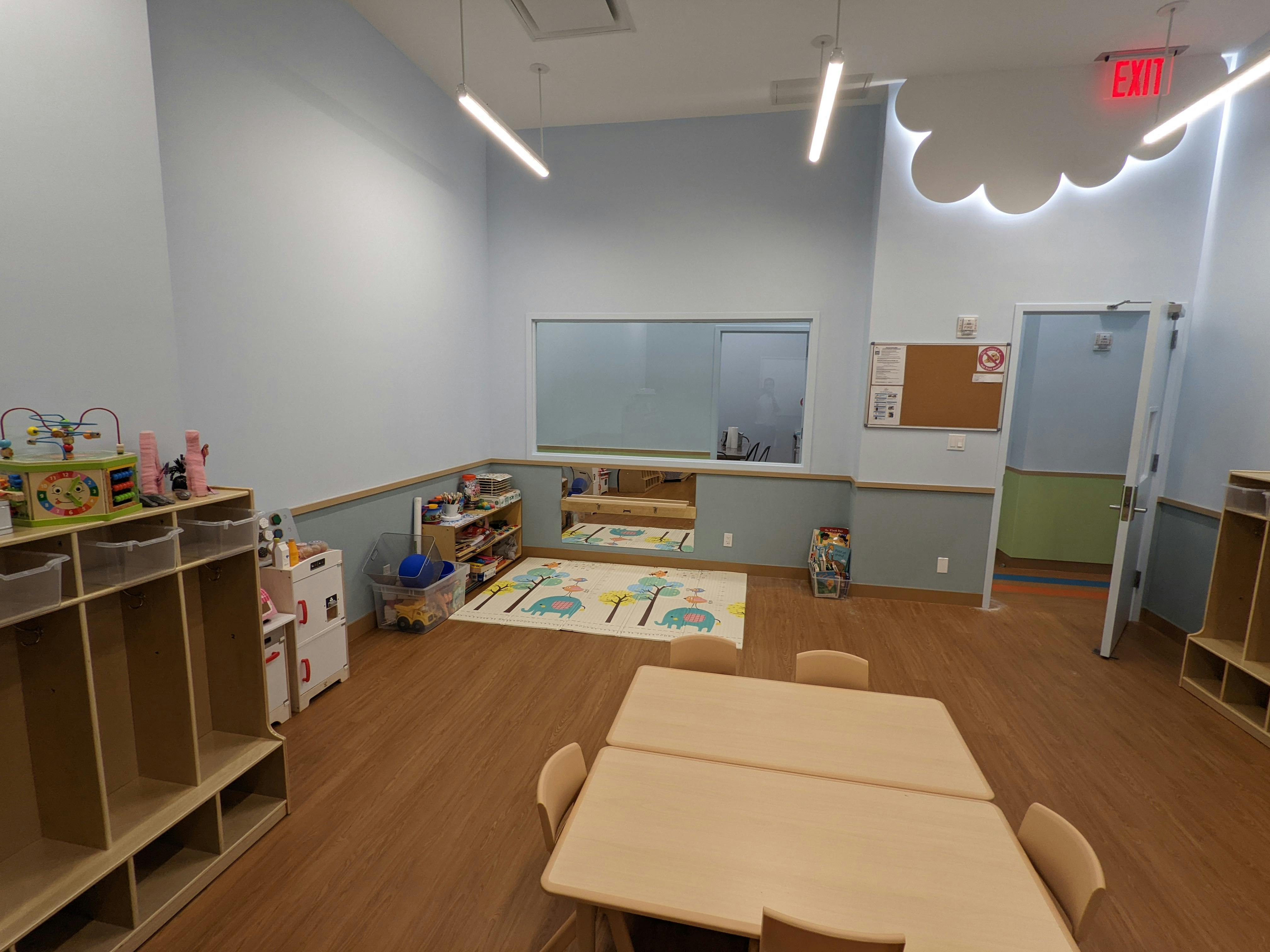 Empty classroom with small tables and chairs, storage shelves, and a play rug