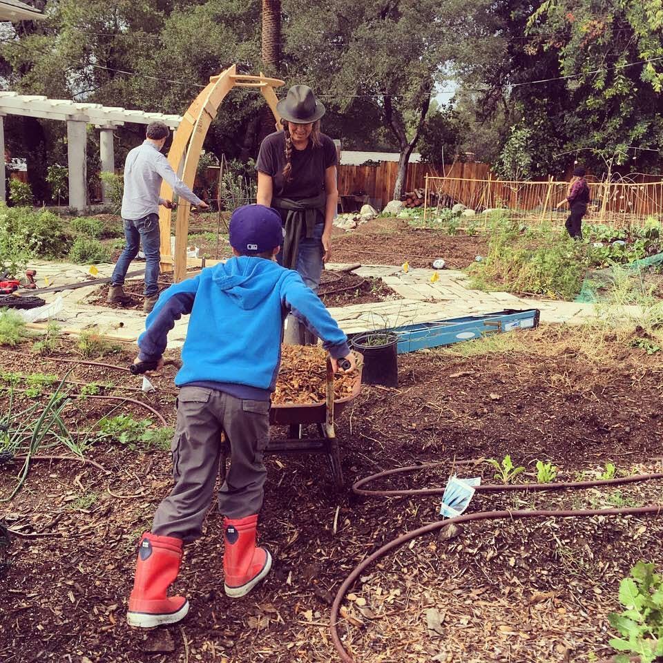Child in red boots pushing a wheelbarrow with a person standing nearby