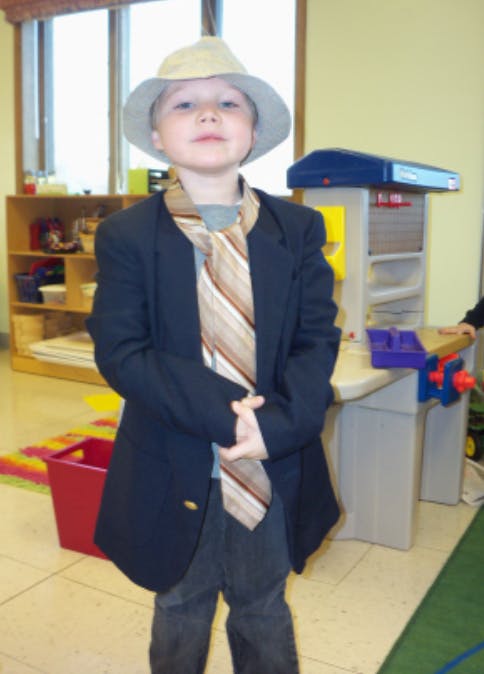 Child wearing an oversized jacket, tie, and hat in a classroom