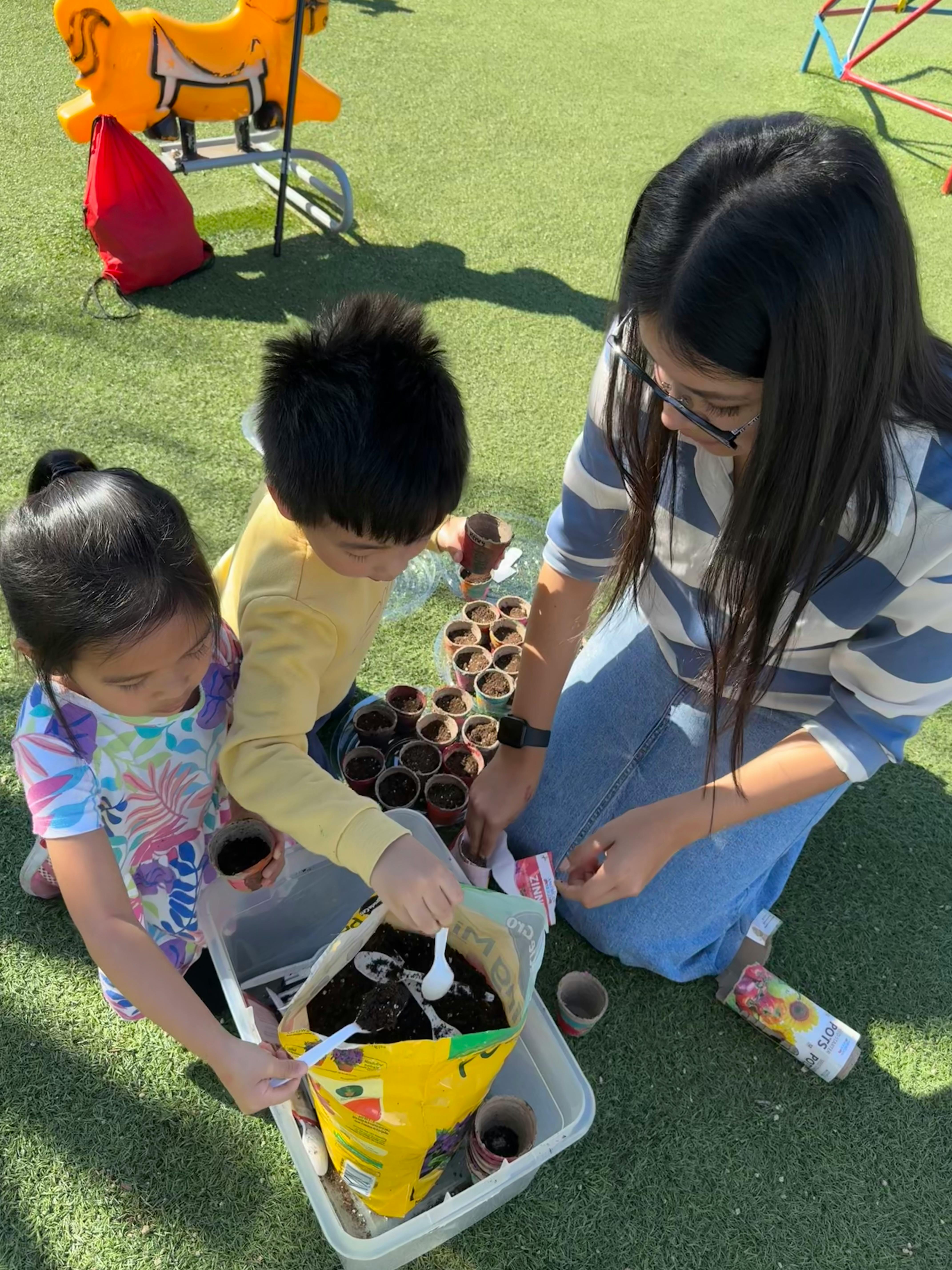 A person and two children planting seeds in small pots outdoors
