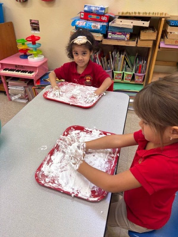 Two children playing with shaving cream on red trays in a classroom