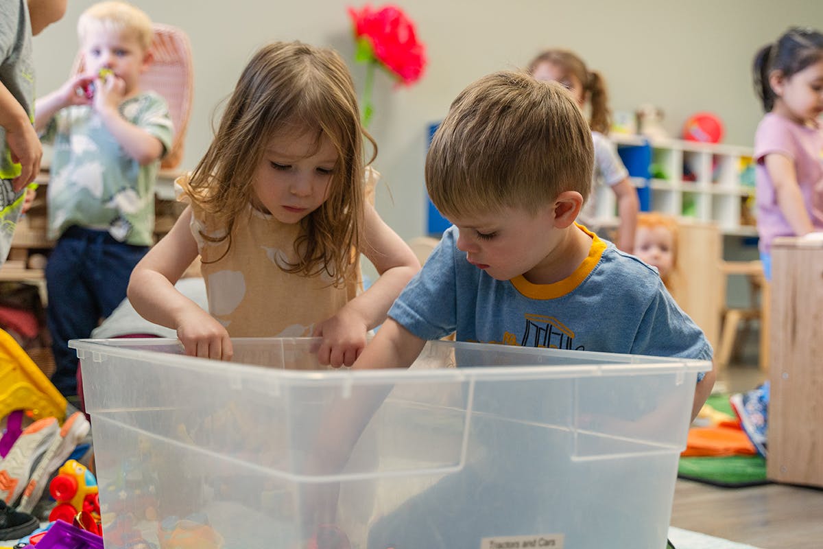 Two children playing with items in a clear plastic bin in a classroom