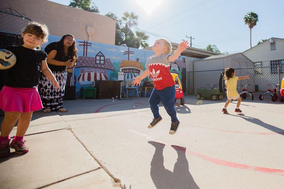 Child jumping in the air outdoors with arms out, other children and adult nearby