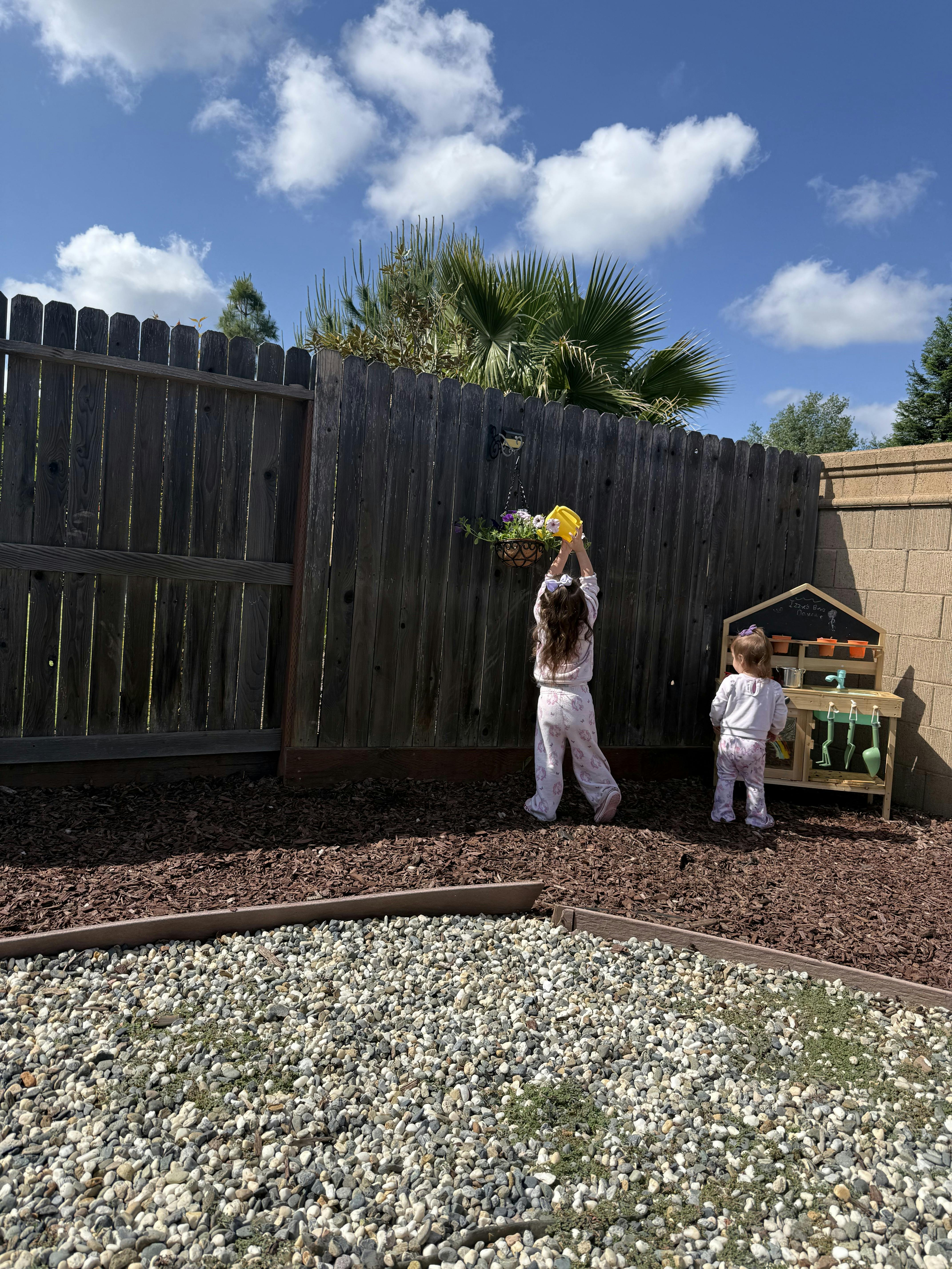 Child watering a hanging flower basket on a wooden fence, another child nearby