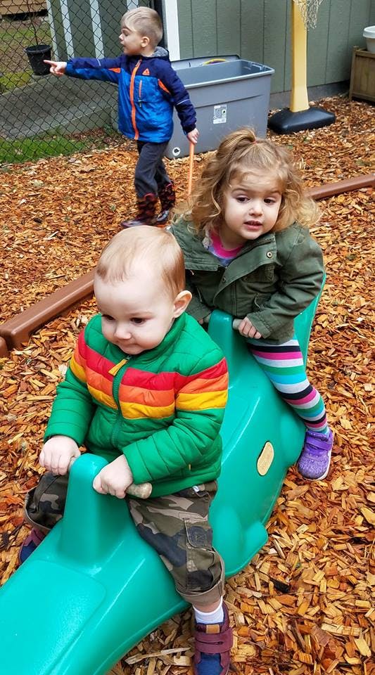 Three children playing on wood chips, two on a green seesaw