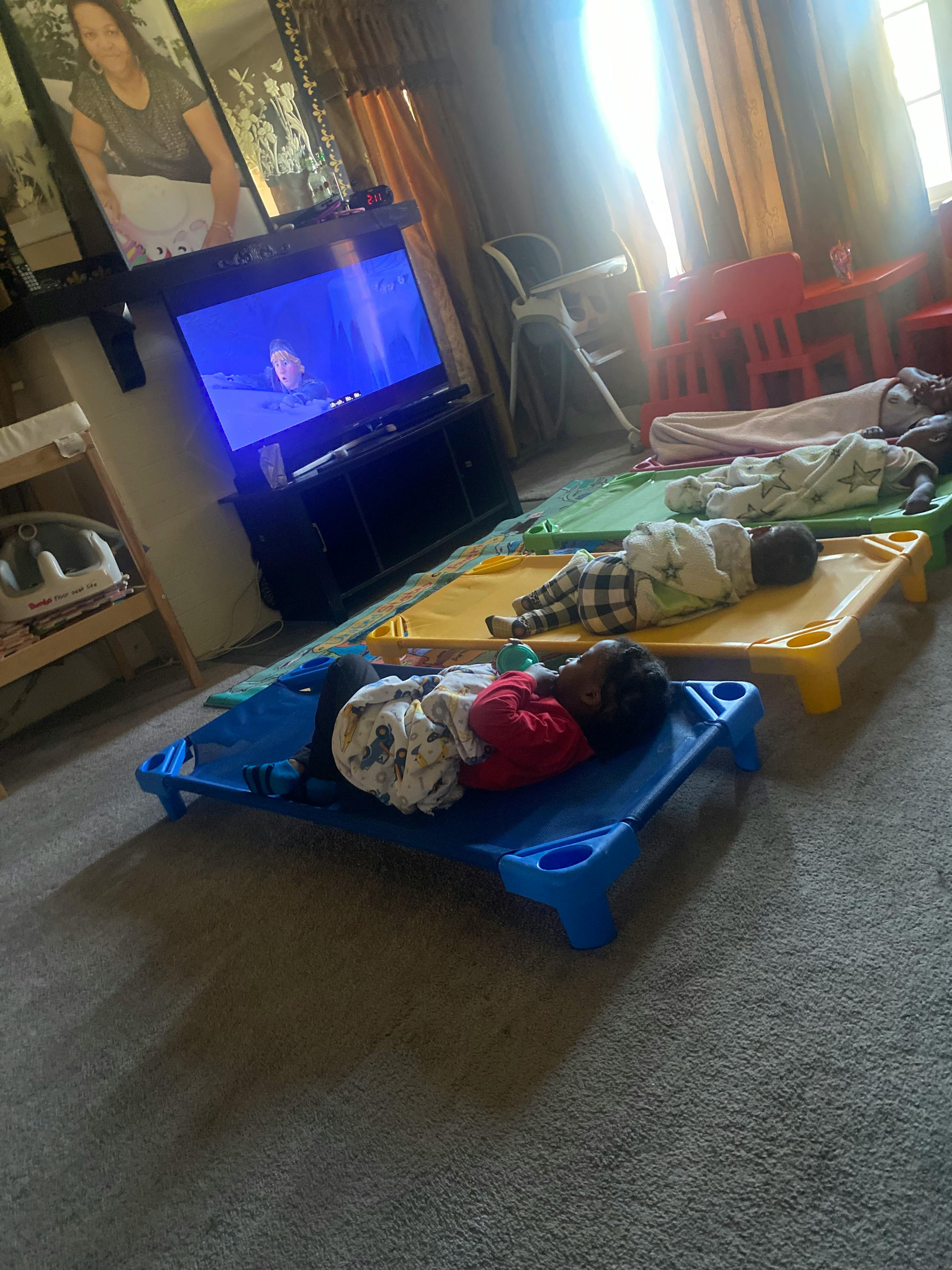 Children resting on cots in a room with a TV playing a movie
