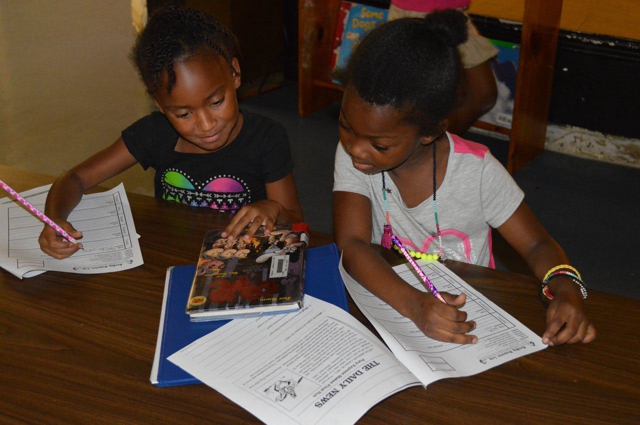 Two children writing at a table with books and papers