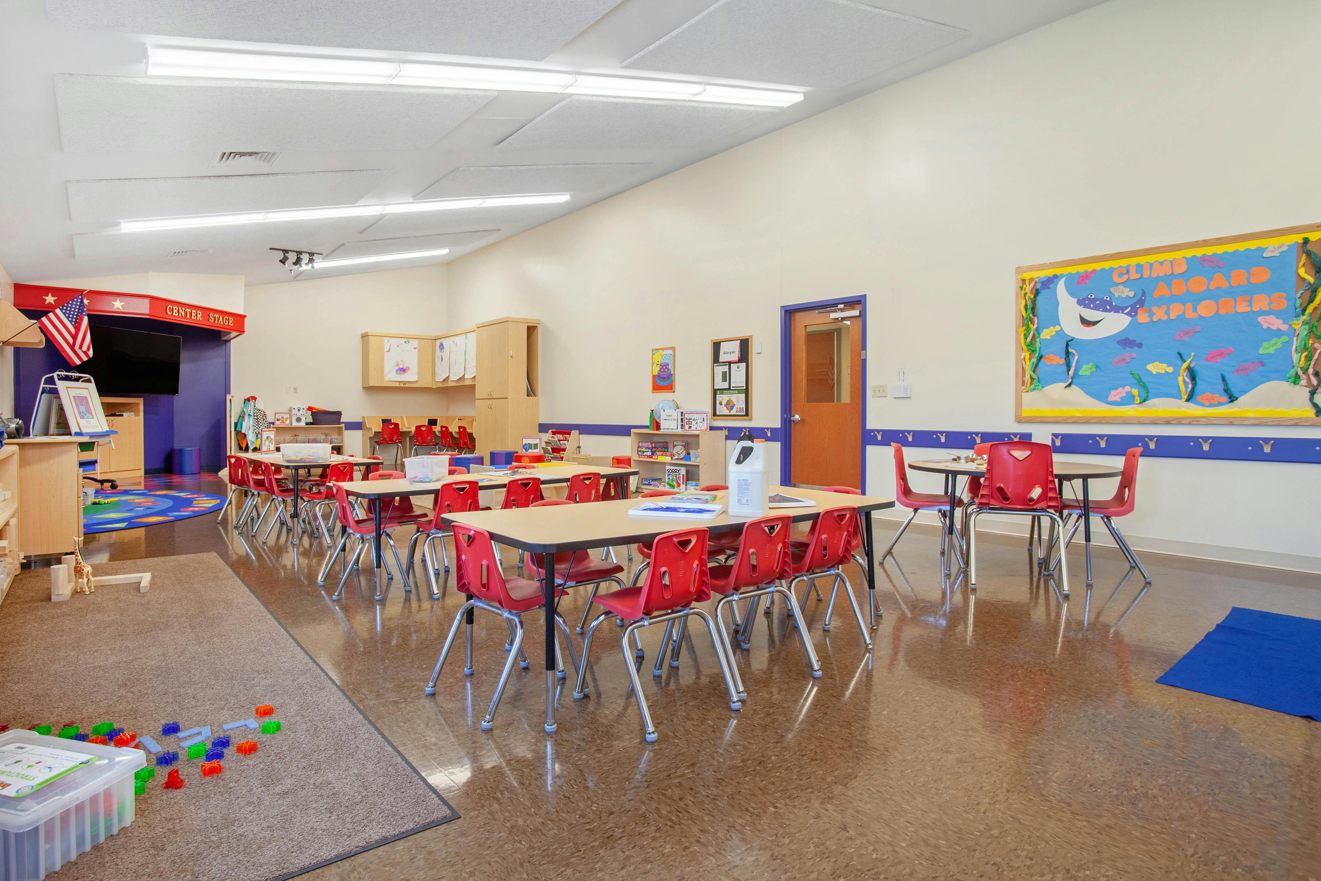 Classroom with small tables and chairs, a stage area, and an 'Explorers' bulletin board
