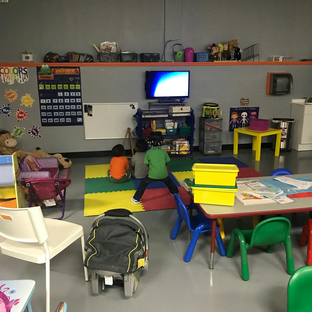 Three children sitting on a colorful mat in a classroom watching television
