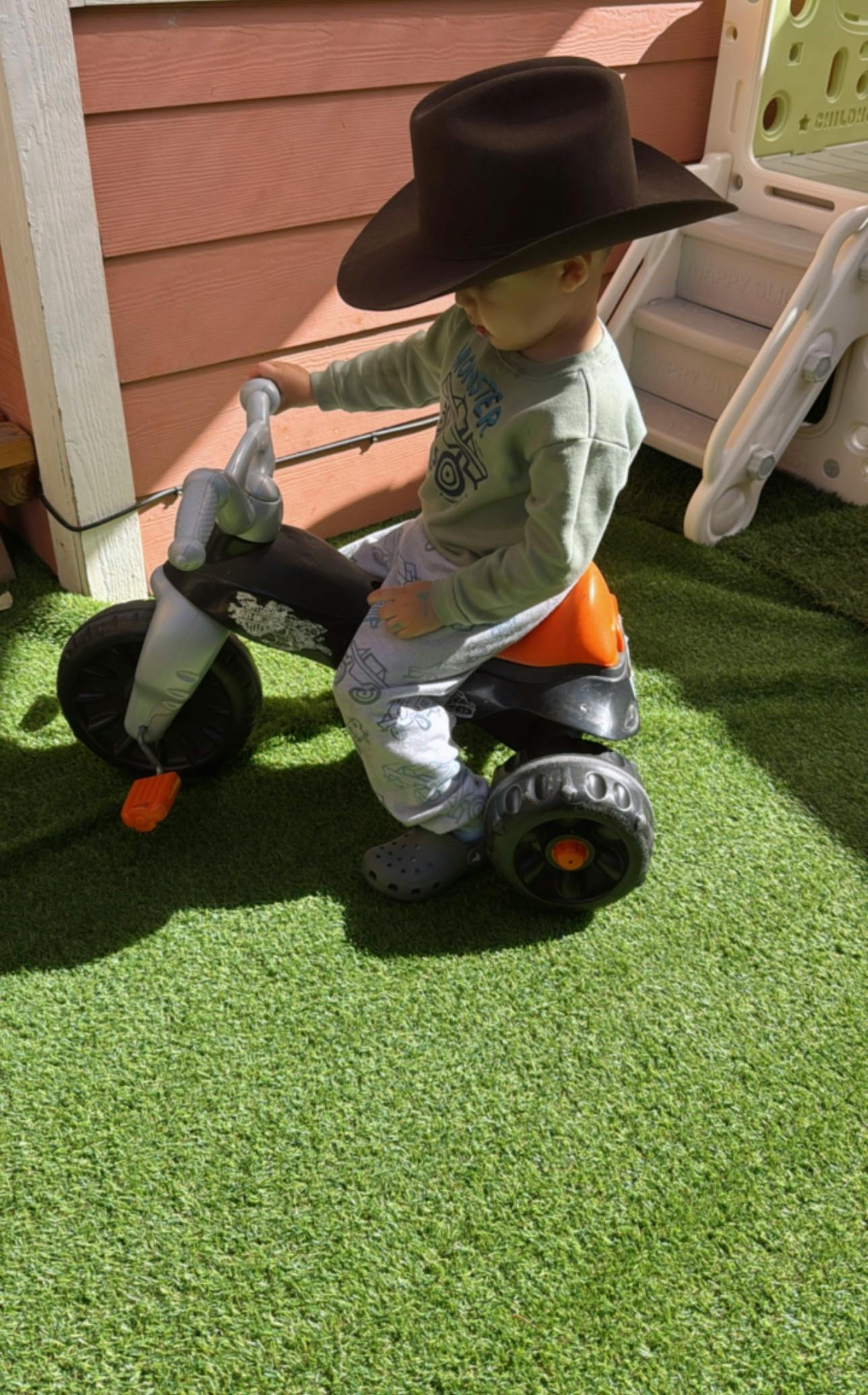 Child wearing a hat riding a toy tricycle on artificial grass