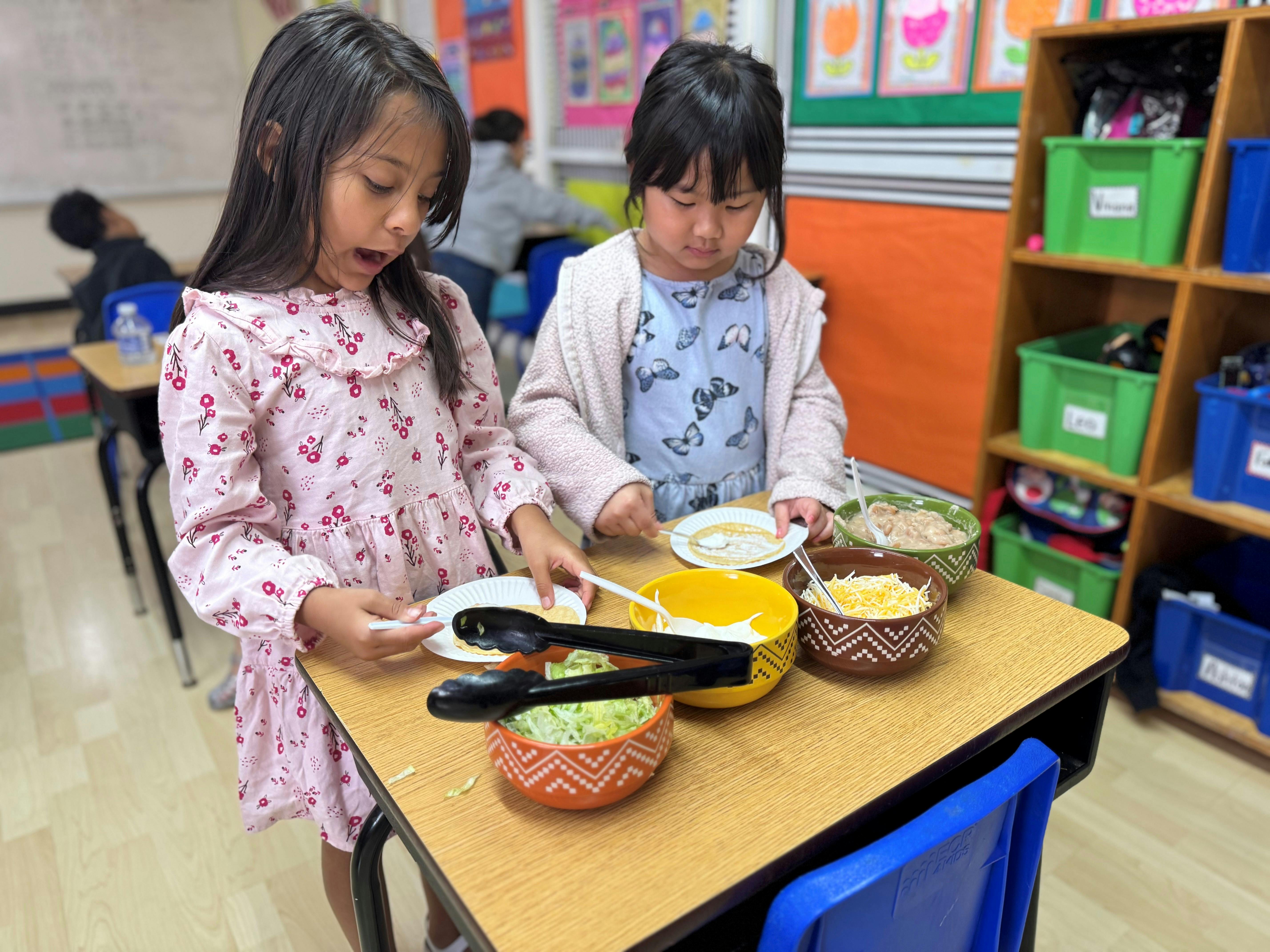 Two children serving food from bowls onto plates in a classroom