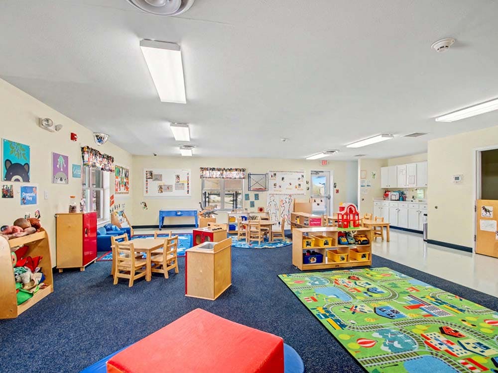 A classroom with tables and chairs, shelves with toys, and a colorful rug