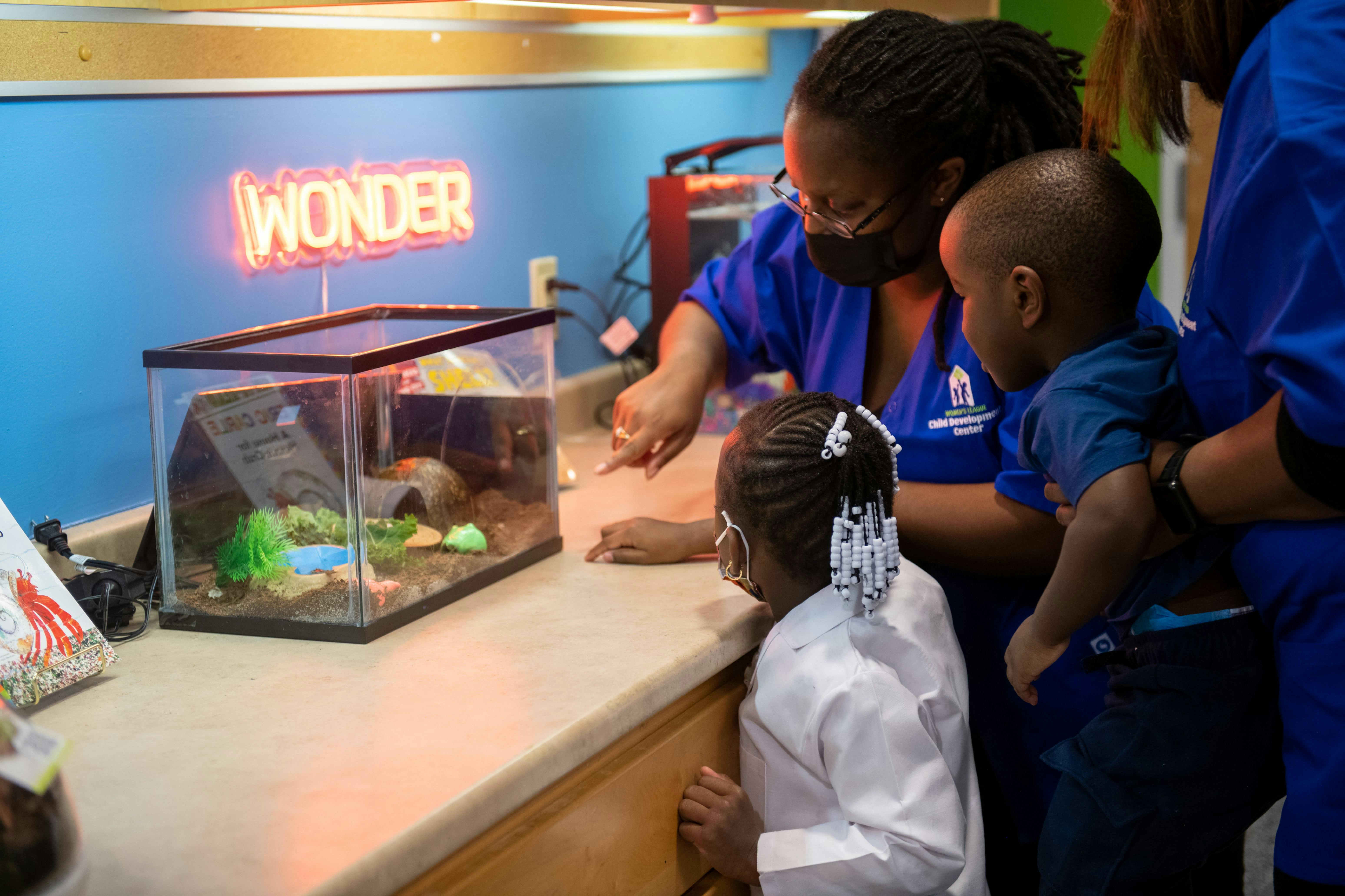 Person pointing at a small animal enclosure with two children looking on a child looking on a baby watching