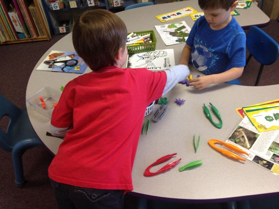Two children playing with small objects and tongs at a table