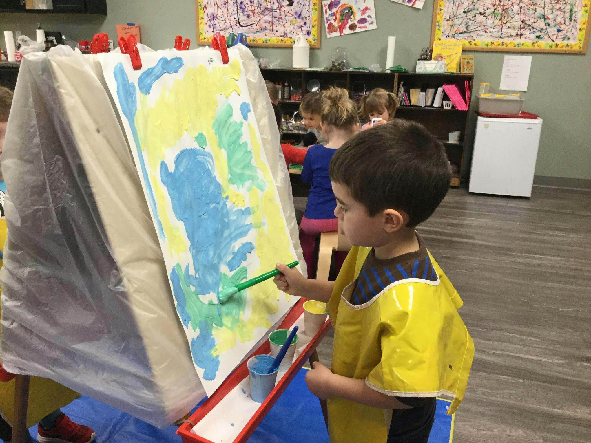 Child in a yellow smock painting on an easel in a classroom