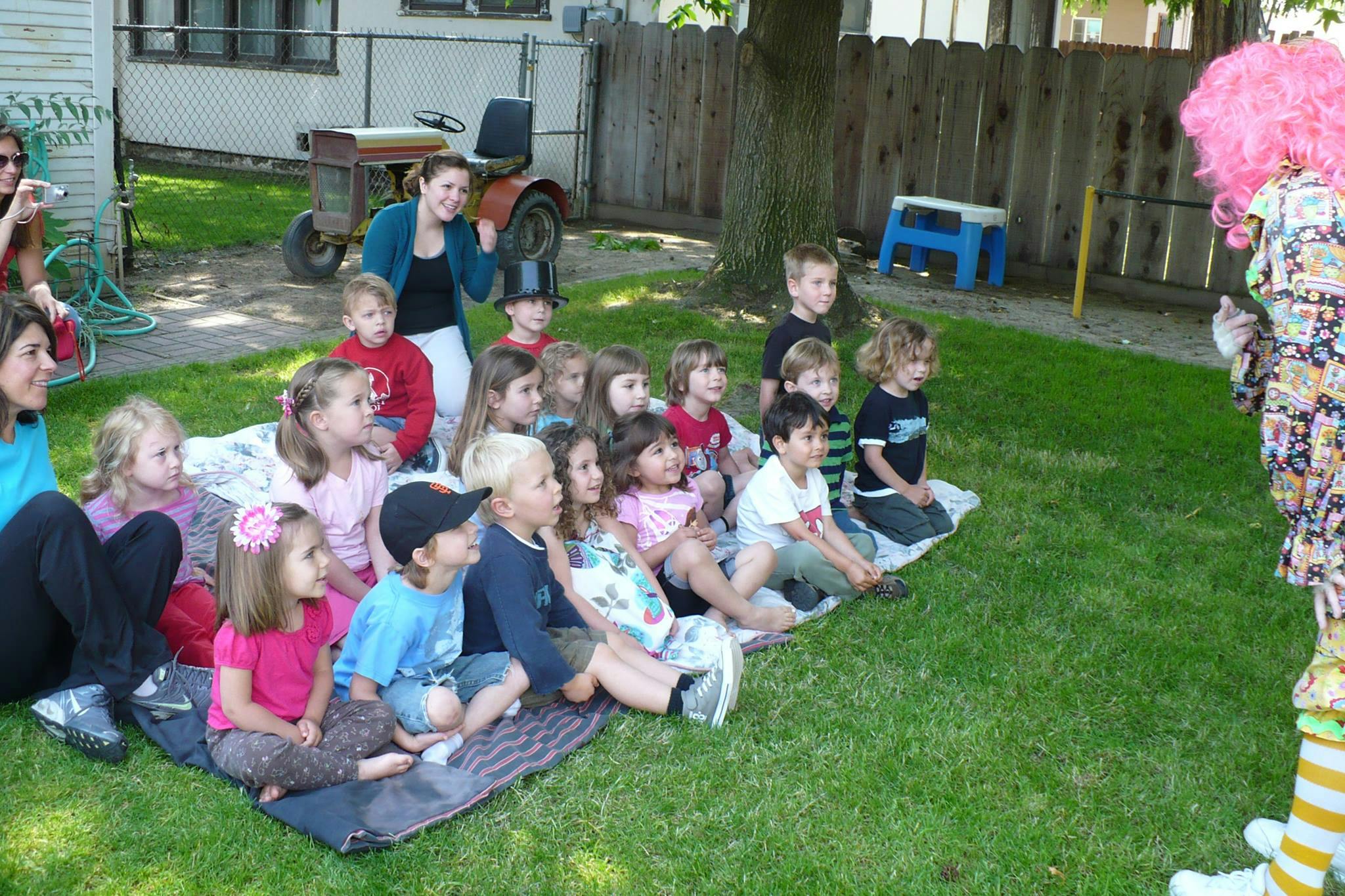 Children and adults sitting on grass watching a person in a costume