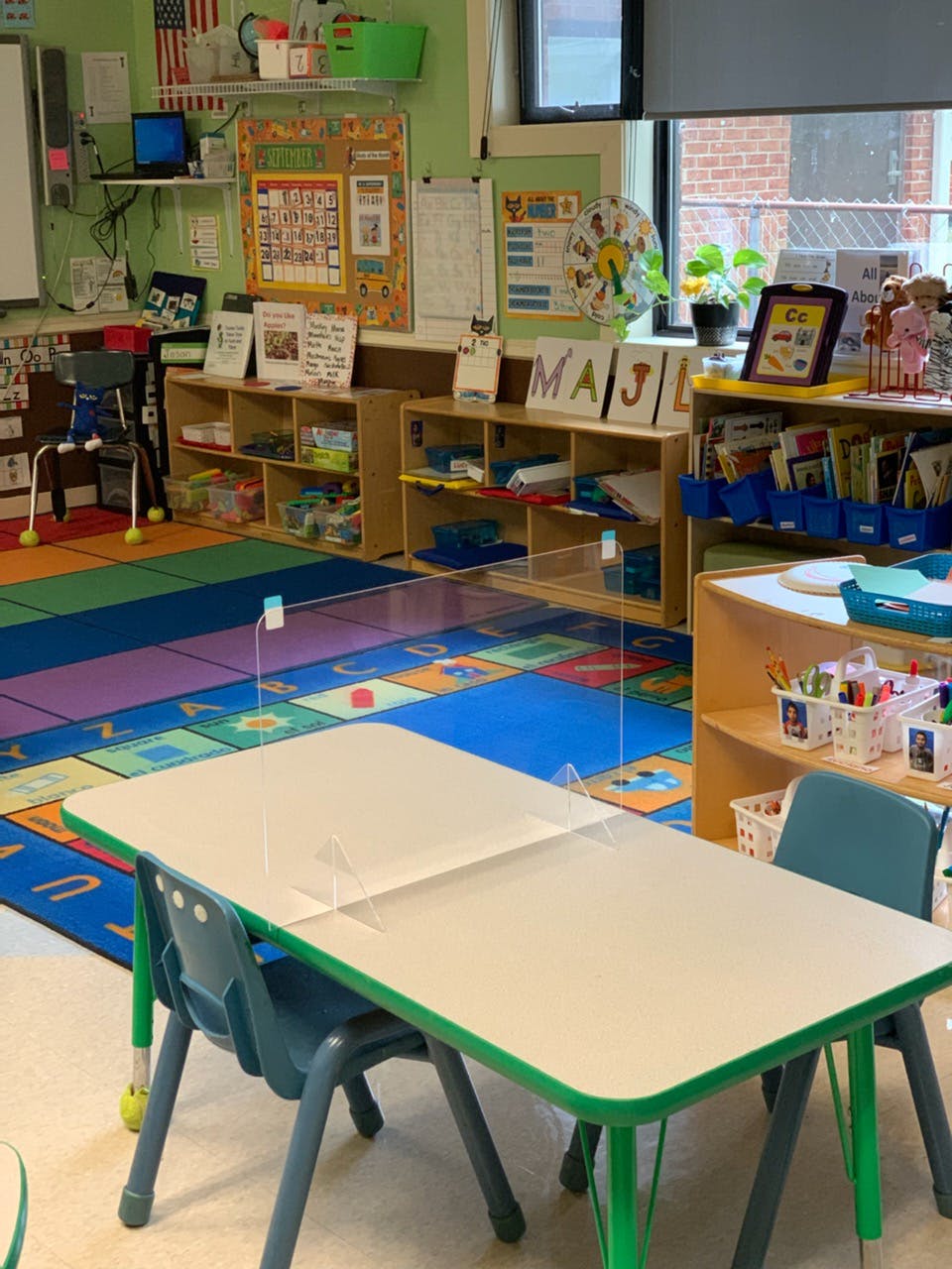 Classroom with small tables and chairs, colorful rug, and shelves with learning materials