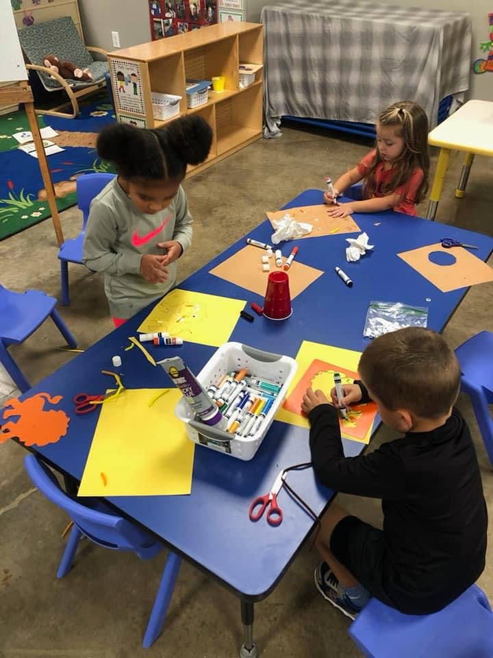 Three children doing crafts at a blue table in a classroom
