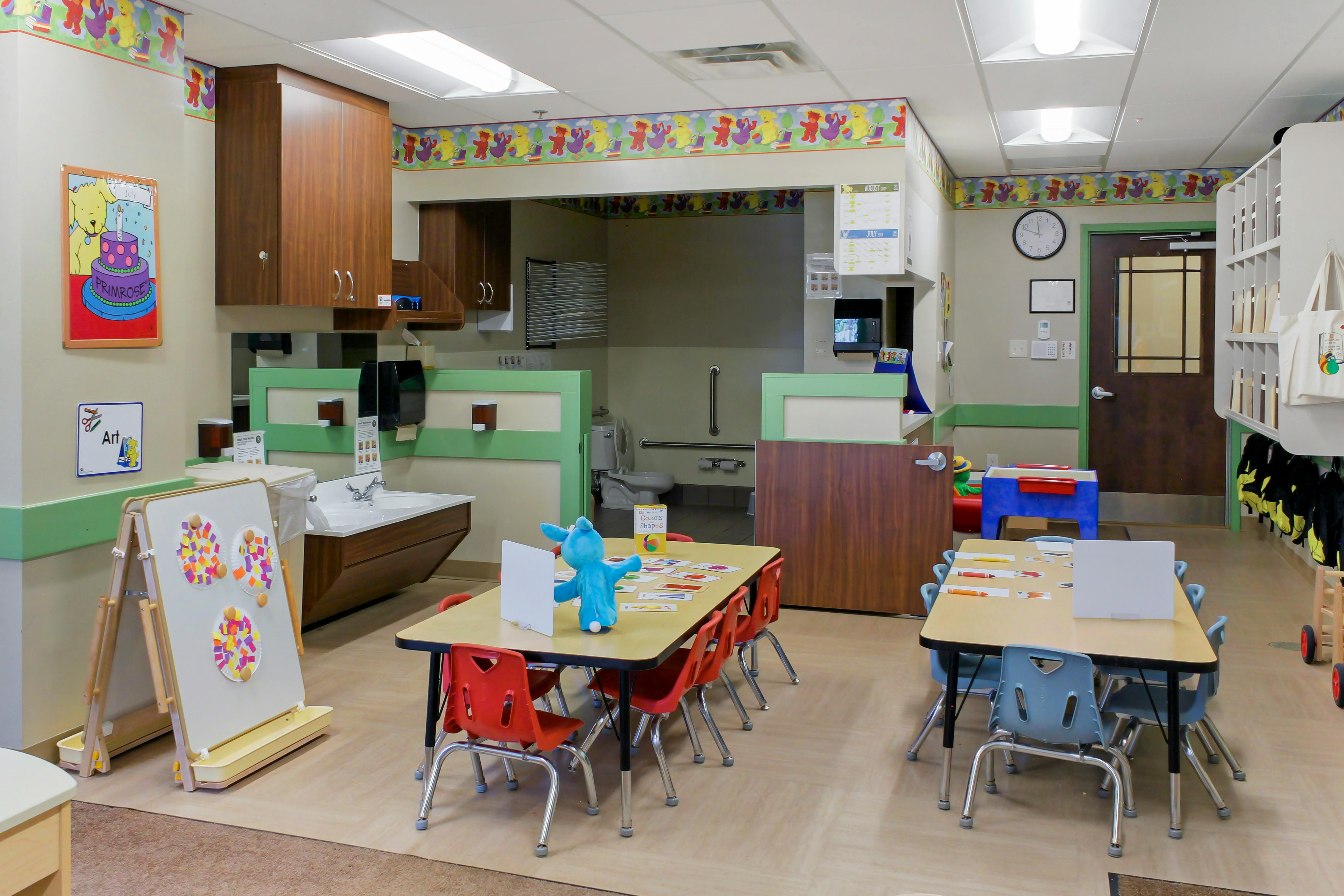 An empty childcare classroom with tables, chairs, sinks, and a toilet stall