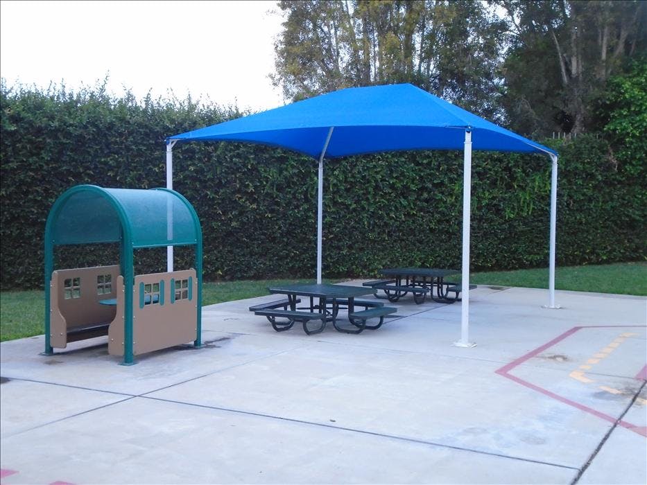Playground area with a blue shade canopy over picnic tables next to a small play structure
