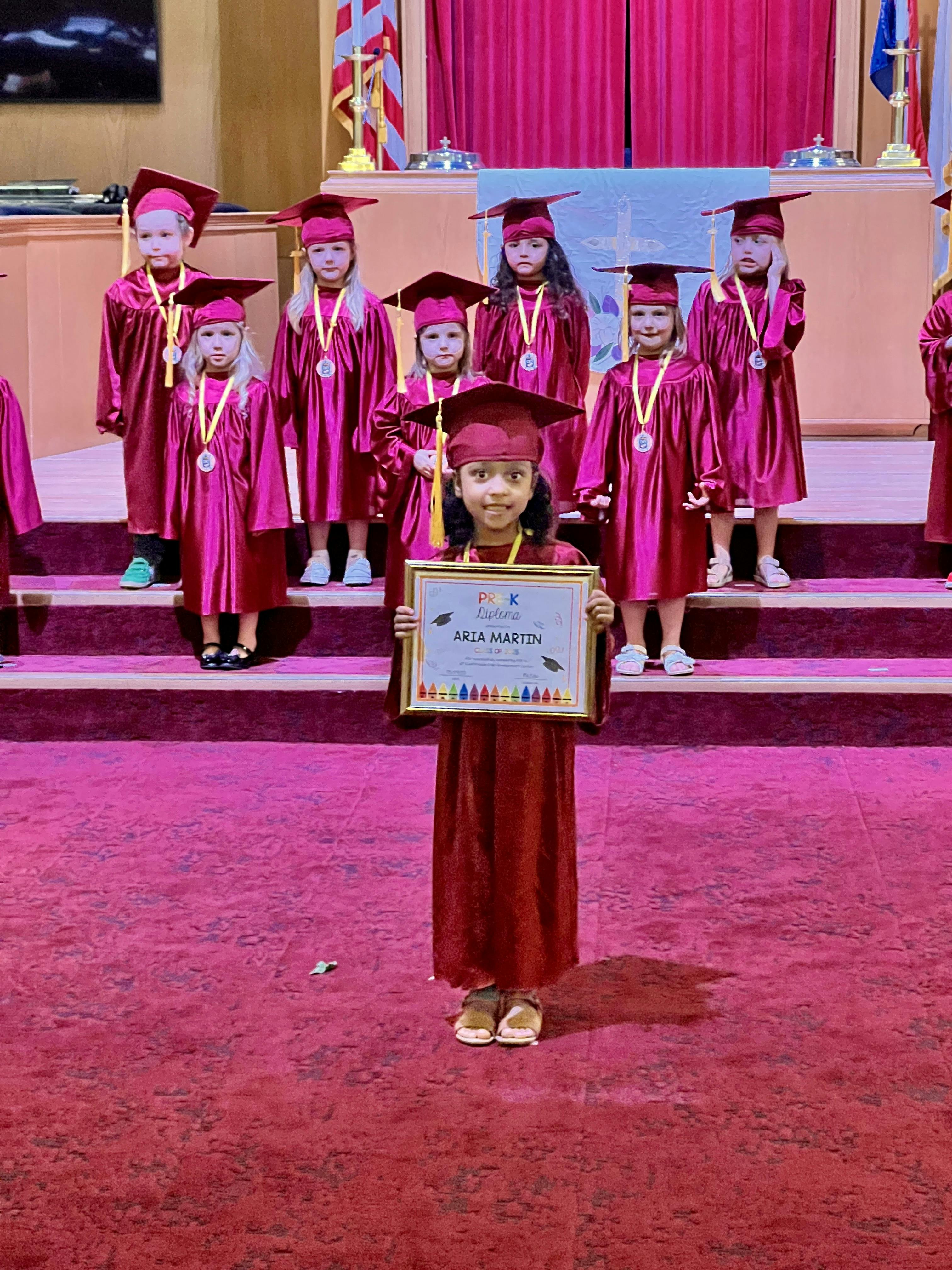 Child in graduation gown holding a framed certificate, other children in background