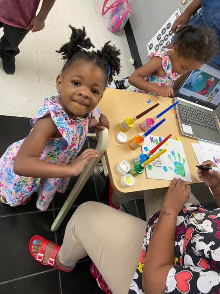 Child looking at camera while another child paints at a table