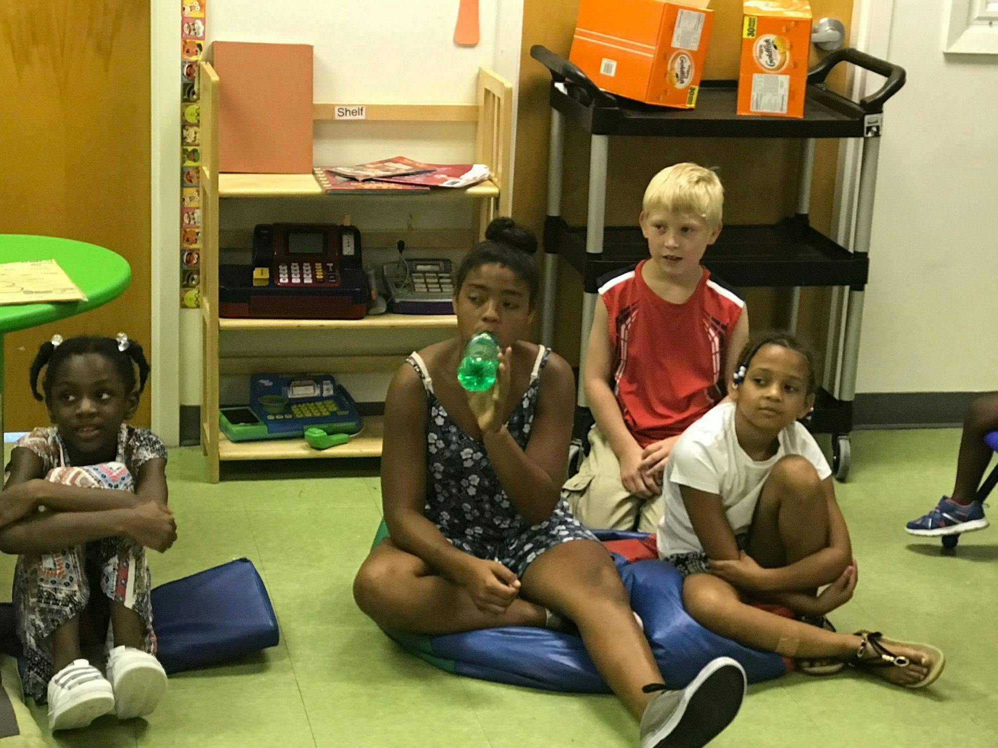 Three children and one teen sitting on the floor, one teen drinking from a bottle