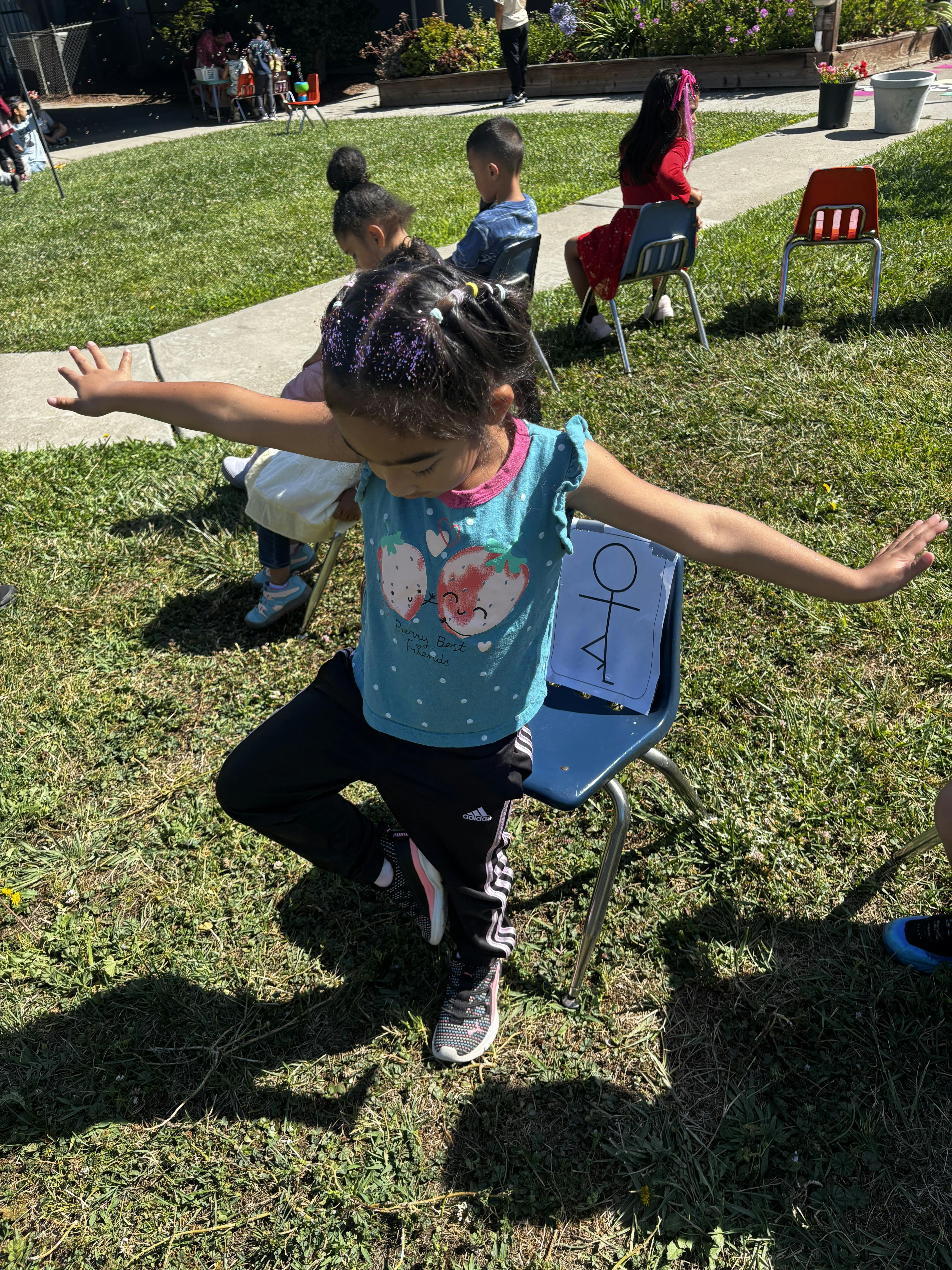 Child balancing on one leg with arms outstretched on a chair outdoors