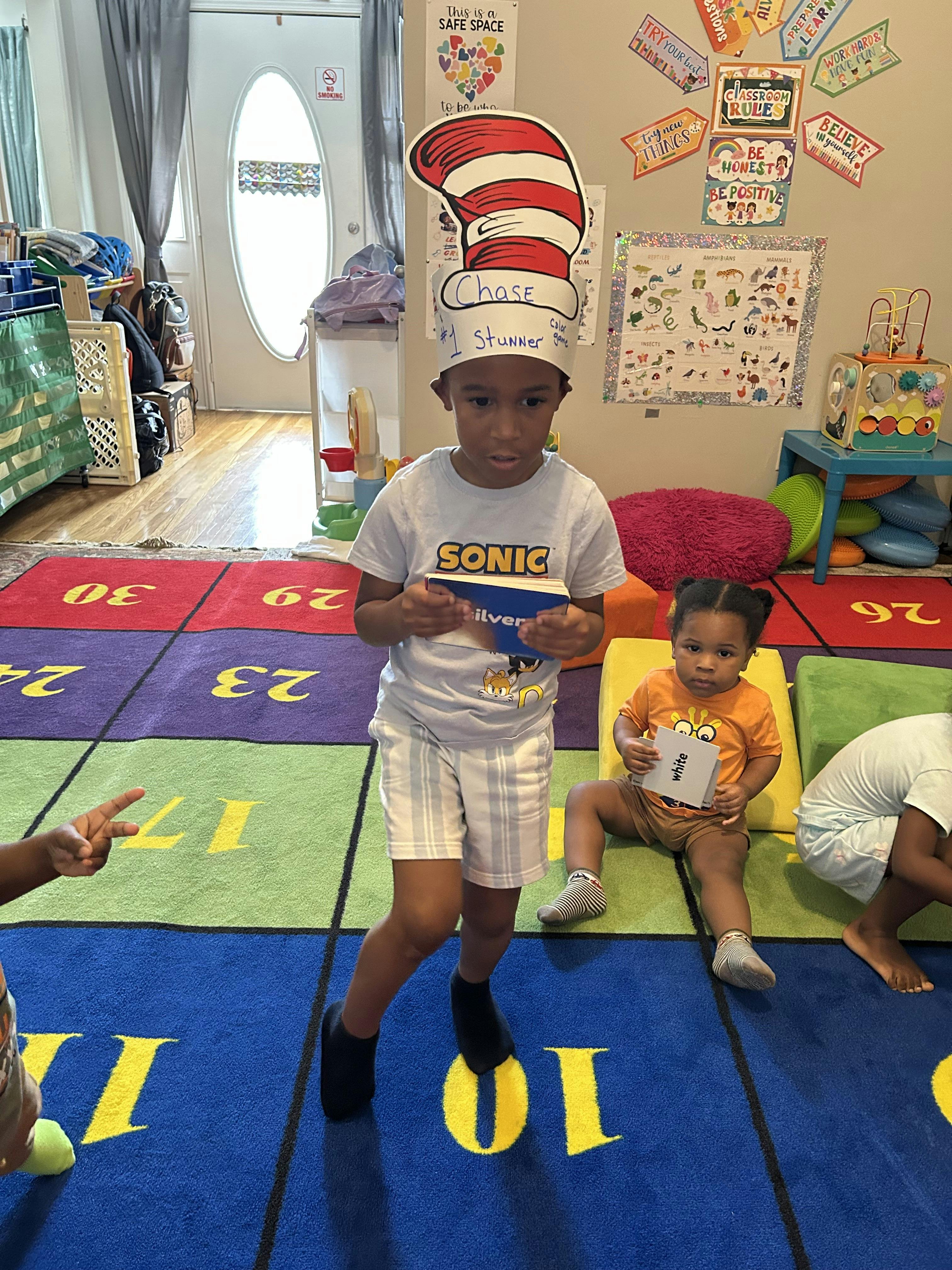 Child wearing a tall striped hat stands in a classroom holding a flashcard
