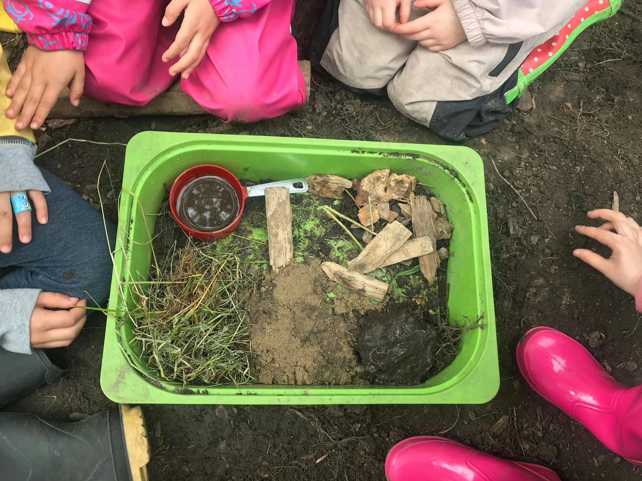 Children kneeling around a green tray with dirt, grass, and wood pieces