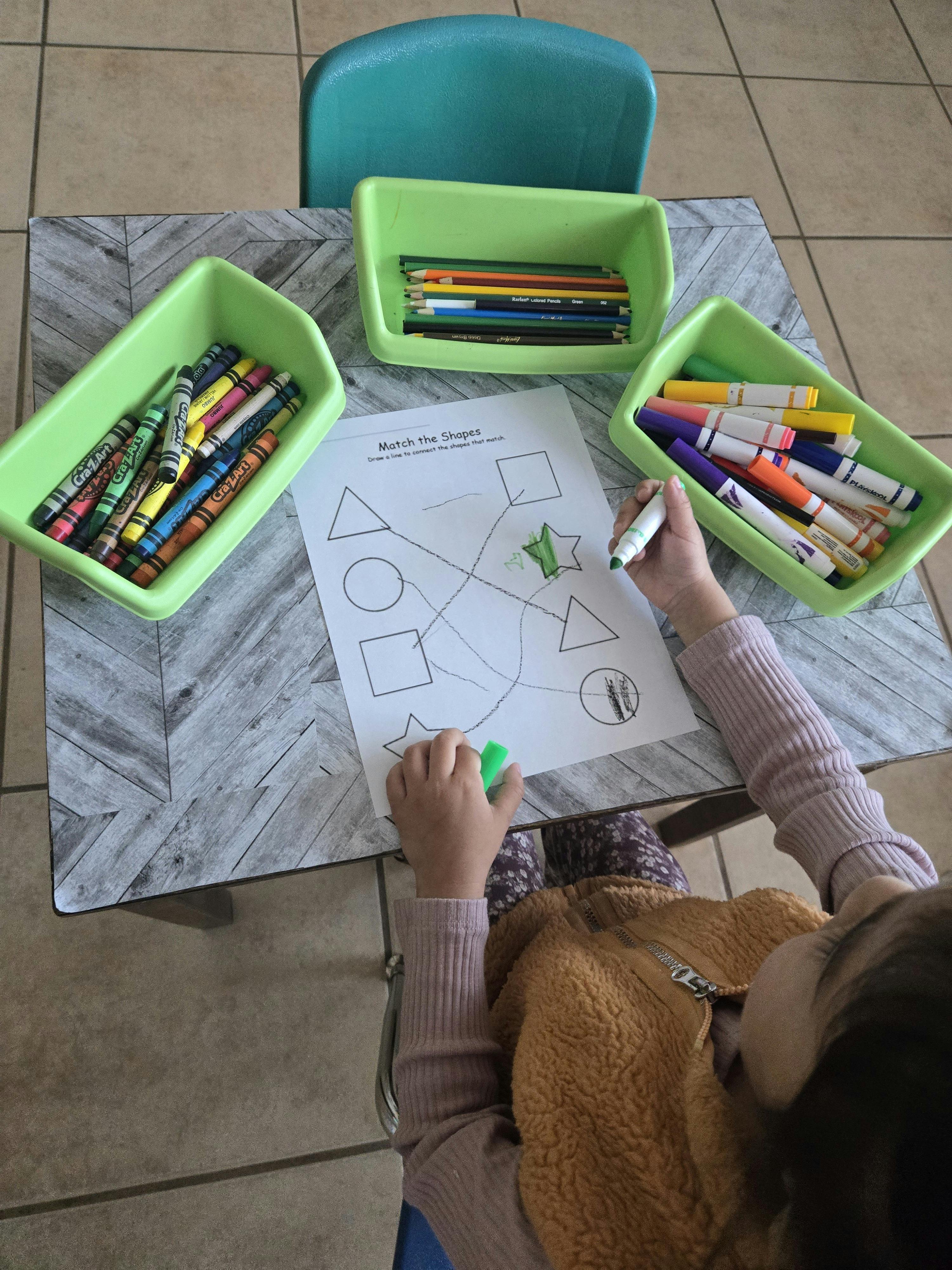 Child matching shapes on a worksheet at a table with crayons and markers