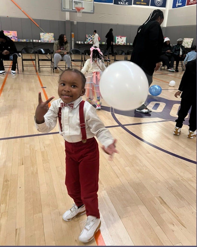 Child in red suspenders and bow tie making a peace sign with a balloon
