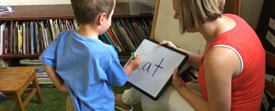Child writing "cat" on a whiteboard held by an adult