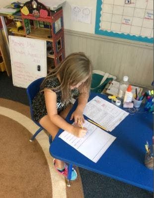 Child writing at a blue table in a classroom