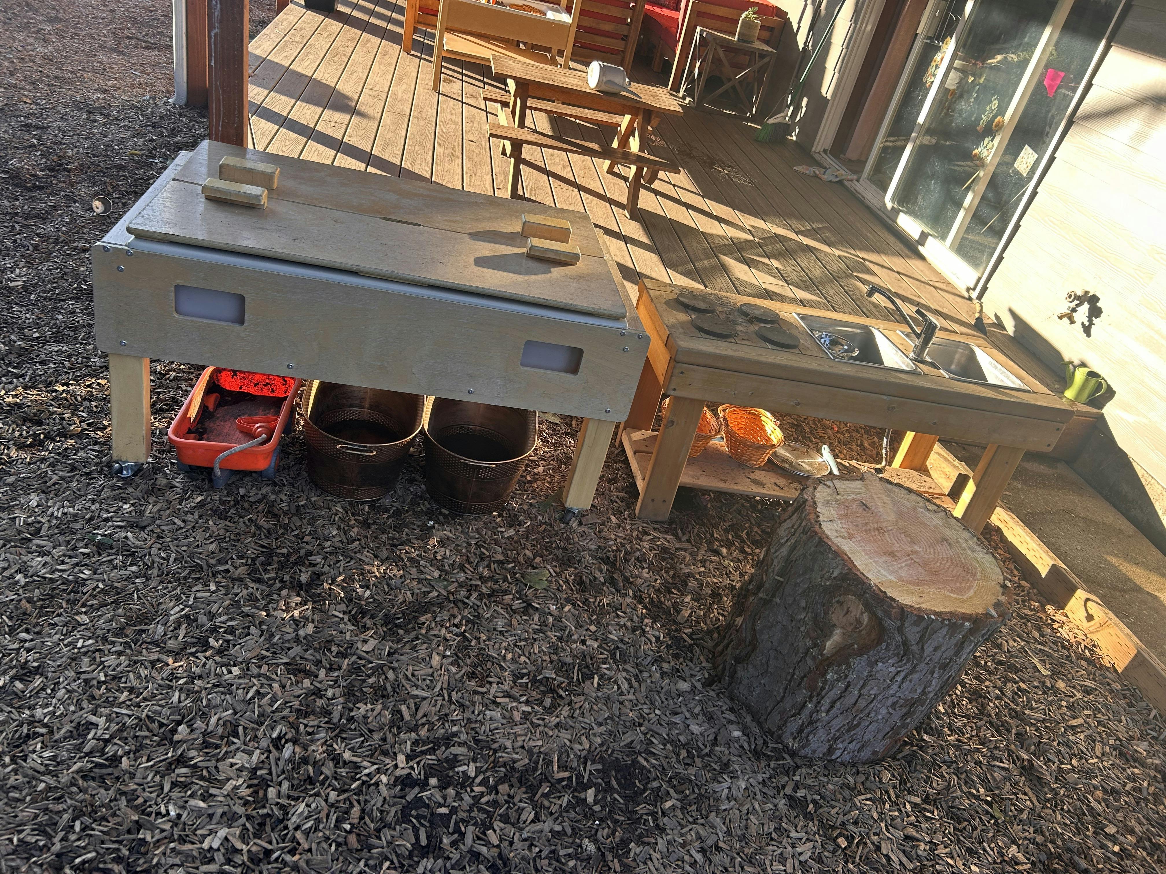 Outdoor play kitchen with two sinks and a mud table on wood chips next to a deck