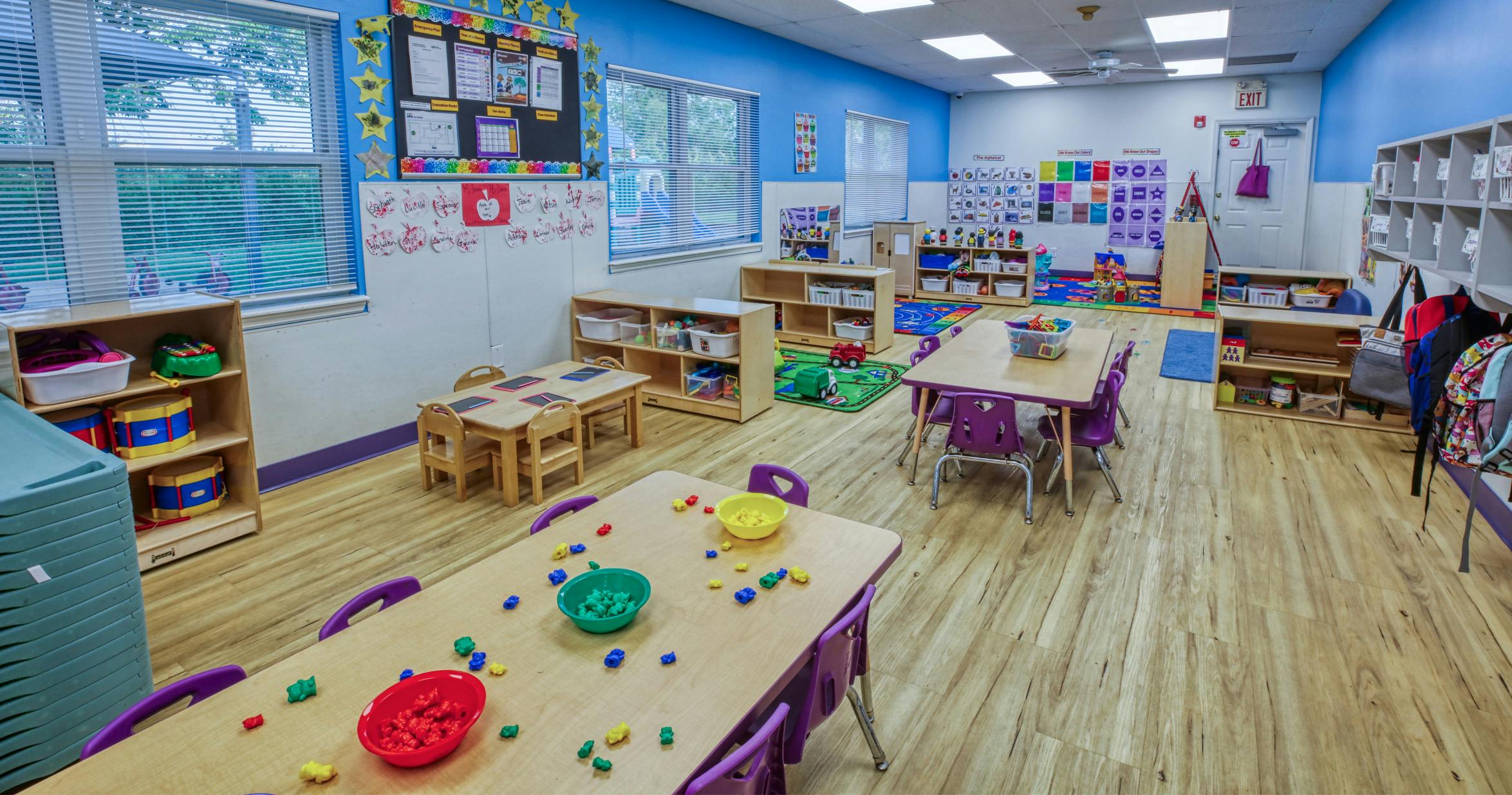 A colorful classroom with small tables and chairs, shelves with toys, and educational posters