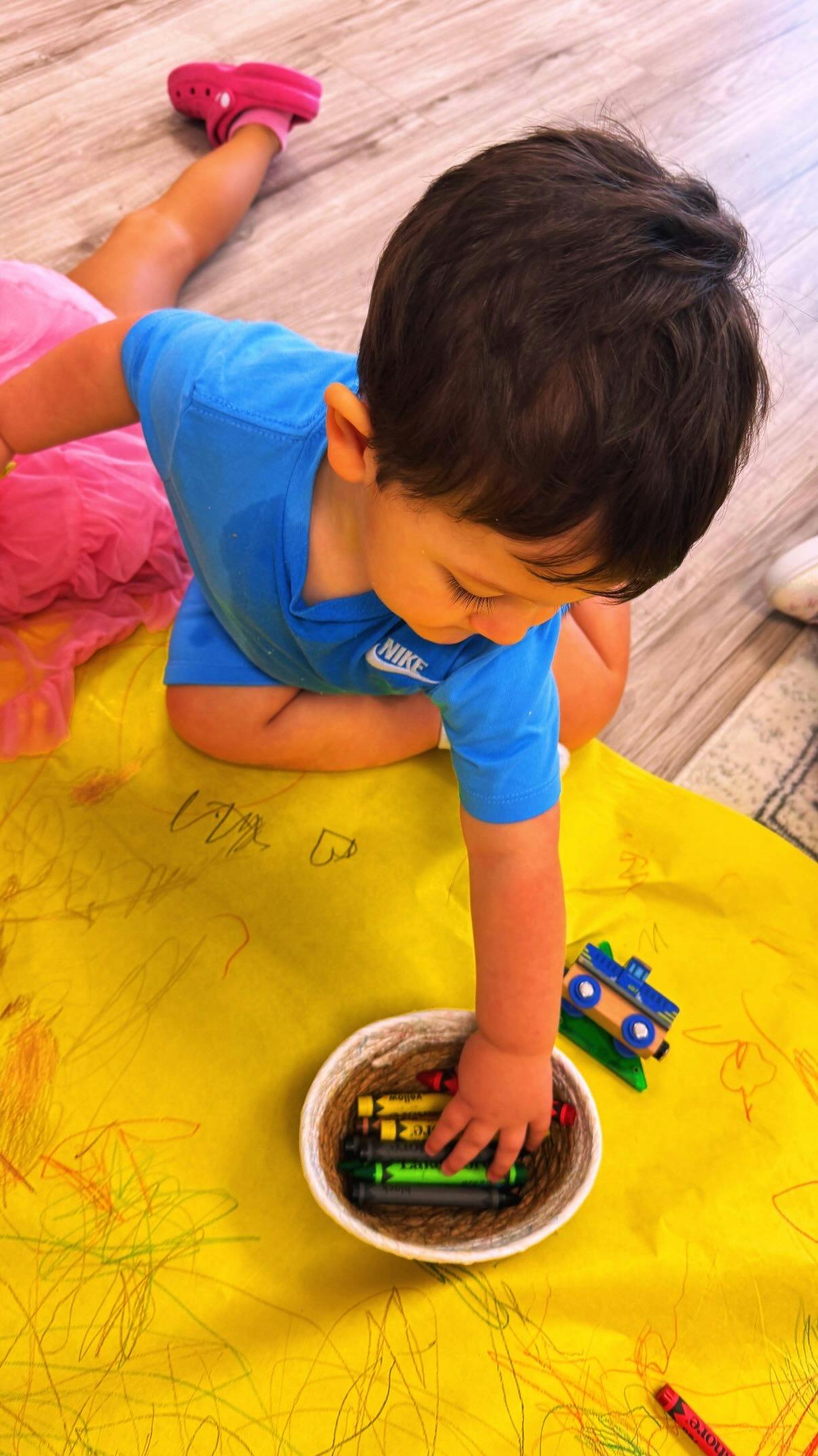 Child reaching for crayons in a bowl on a yellow paper surface