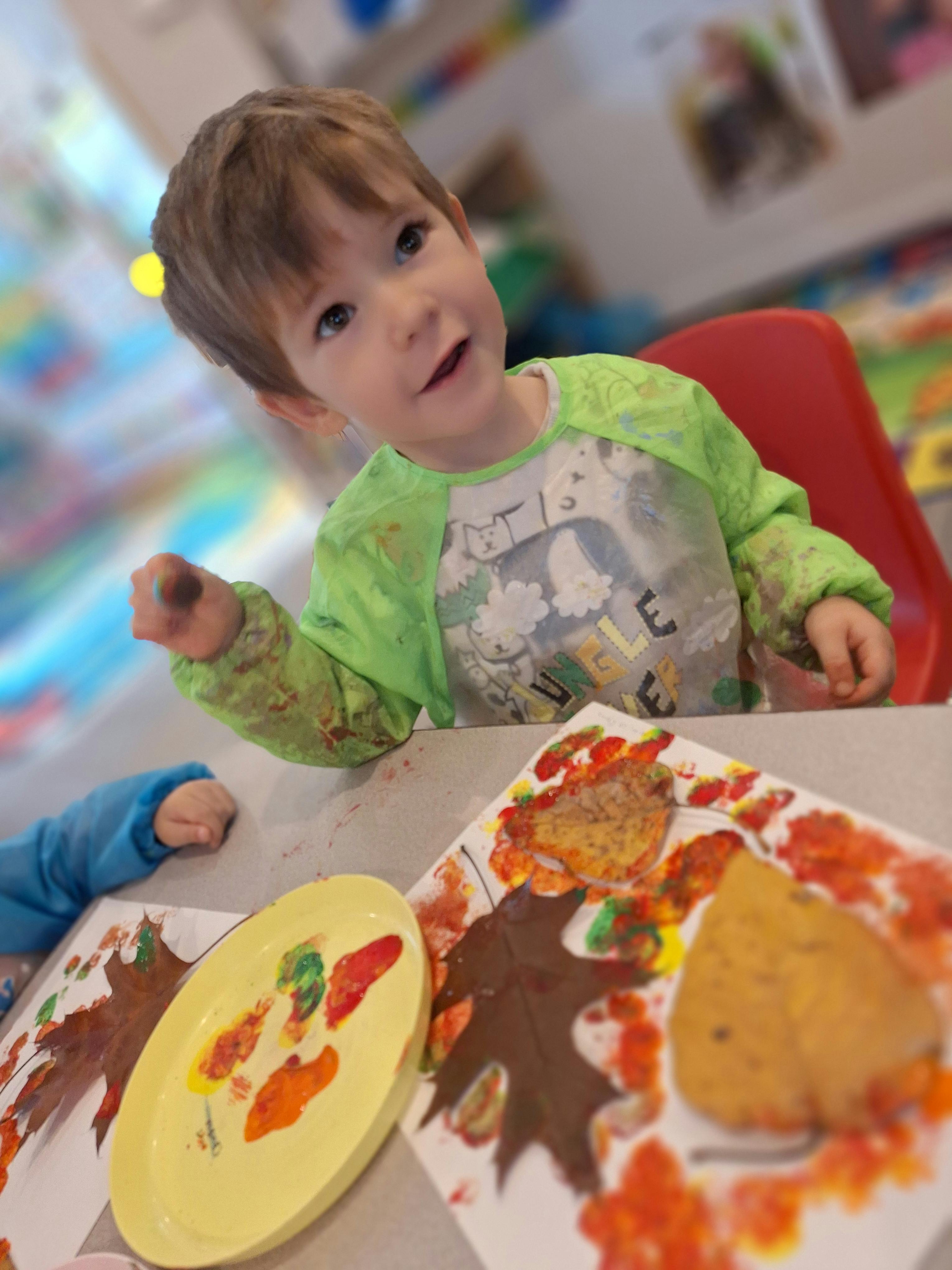 Child wearing a green smock doing a leaf painting craft with red and orange paint