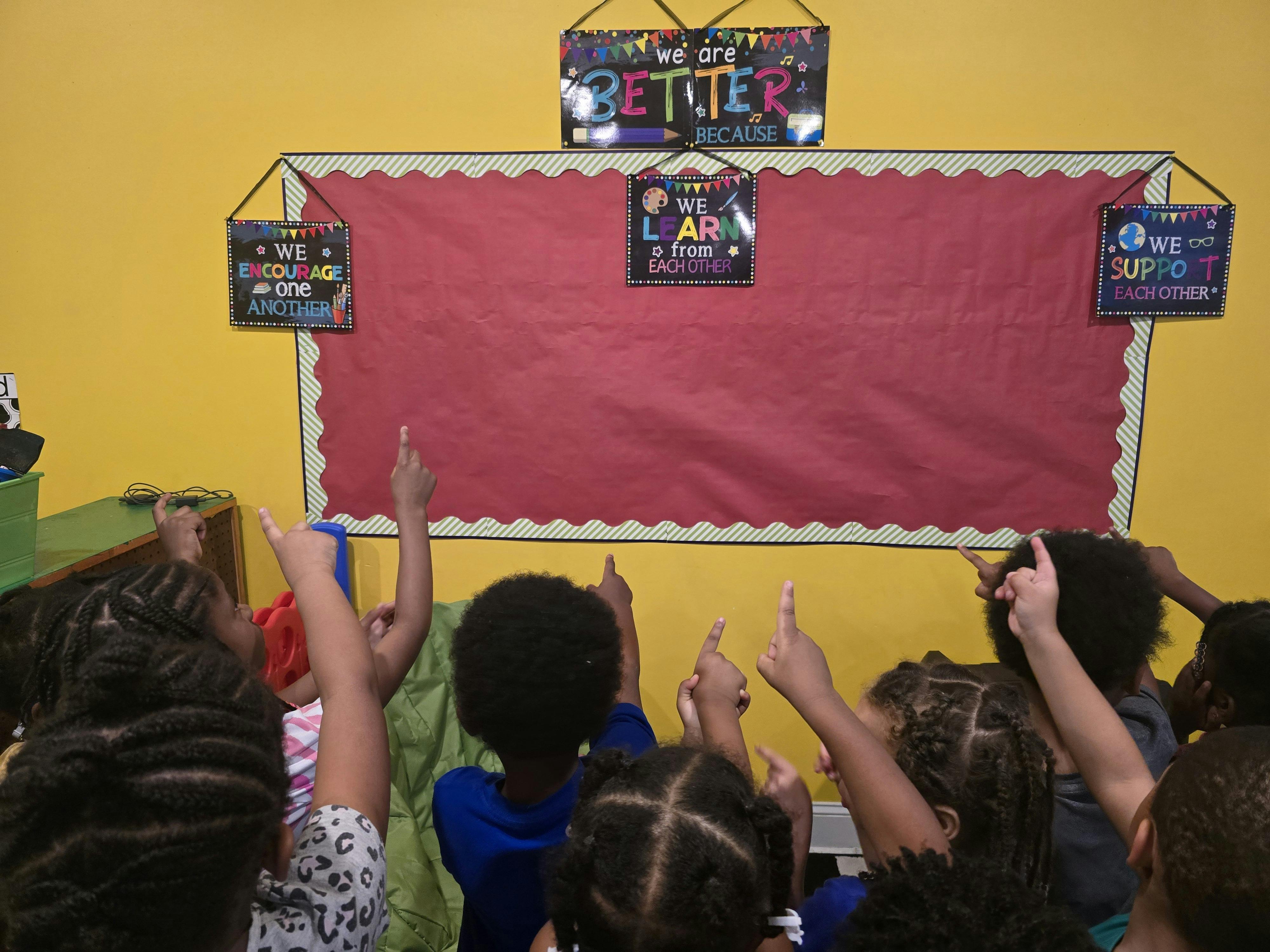 Group of children pointing at a blank bulletin board in a classroom