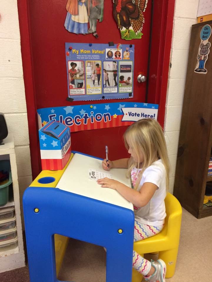 Child sitting at a desk, writing on a paper next to a ballot box