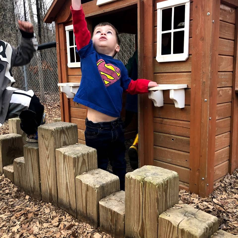 Child in Superman shirt reaching up next to a playhouse
