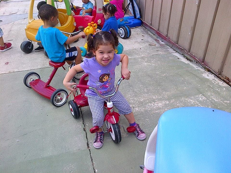 Child on a red tricycle with another child on a scooter in the background