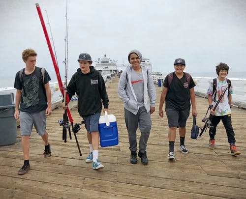 Five teens walking on a pier carrying fishing rods and a cooler